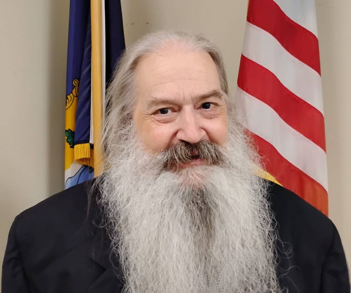 An older man with a long white beard and moustache smiles in front of the American flag and another flag. He wears a black jacket.