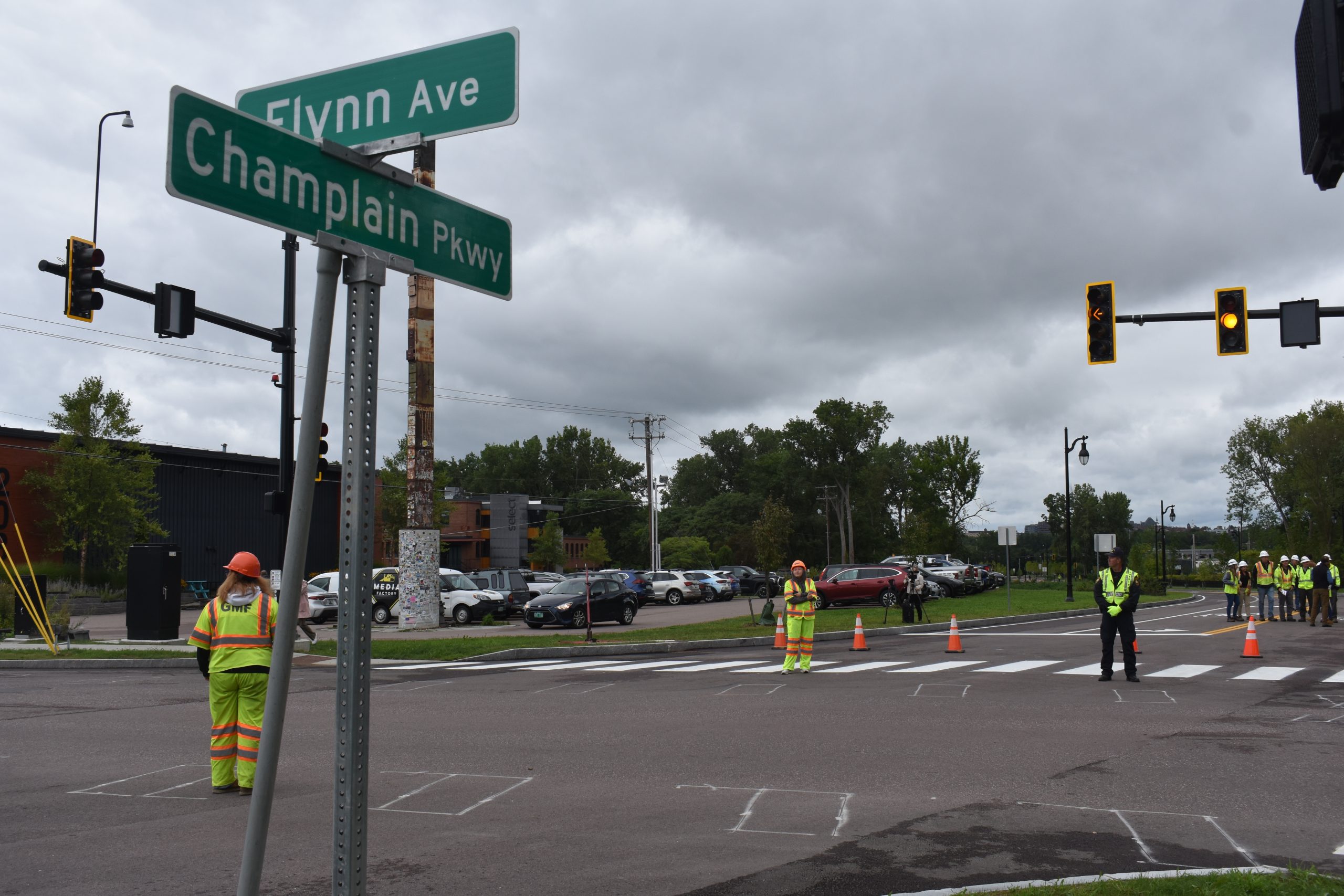 Construction workers in high-visibility vests manage traffic at an intersection with Flynn Ave and Champlain Pkwy signs. Traffic cones are set up, and several workers are near a crosswalk.