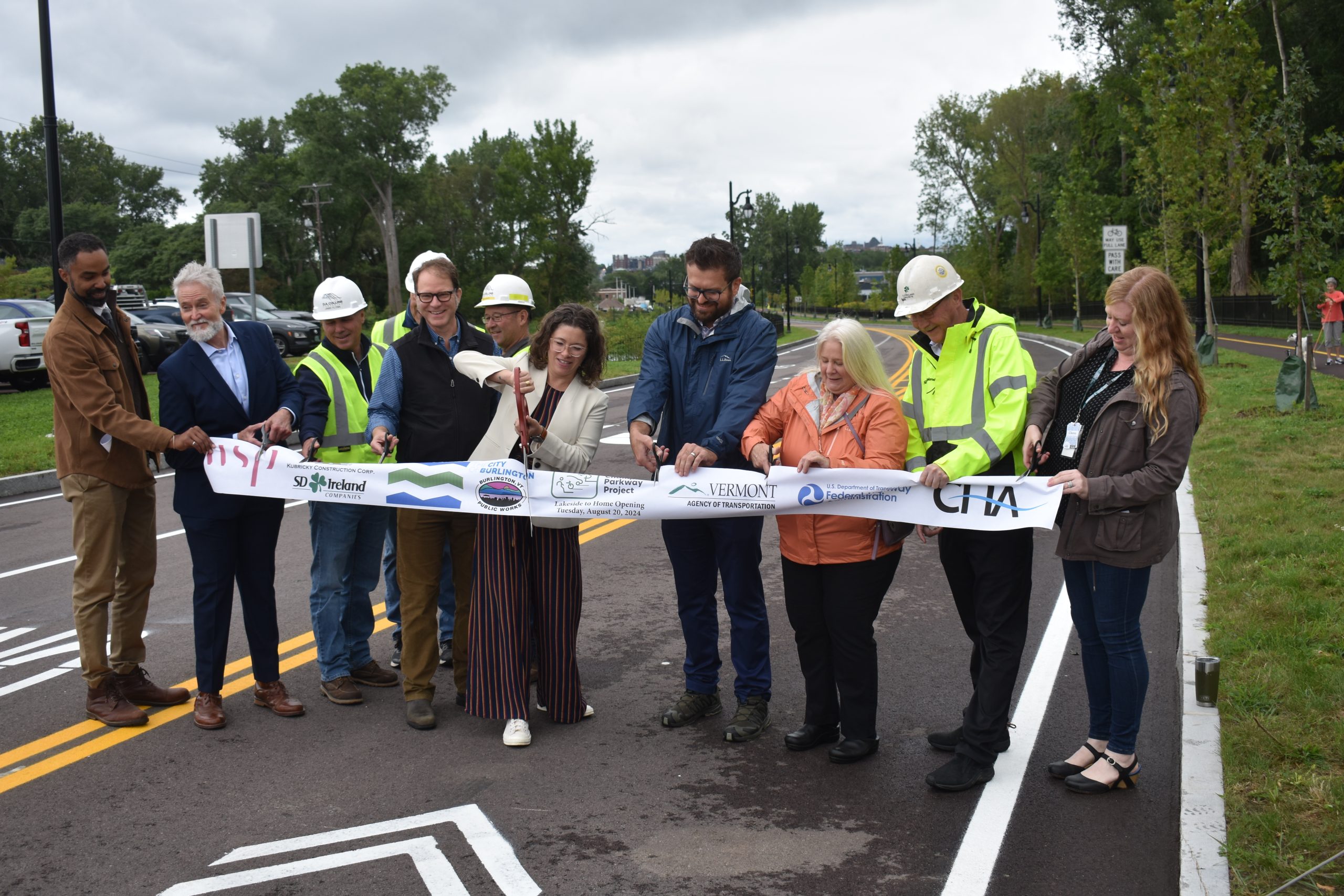 A group of people, including construction workers and officials, stand on a new road cutting a ceremonial ribbon. The event is outdoors with trees and a cloudy sky in the background.