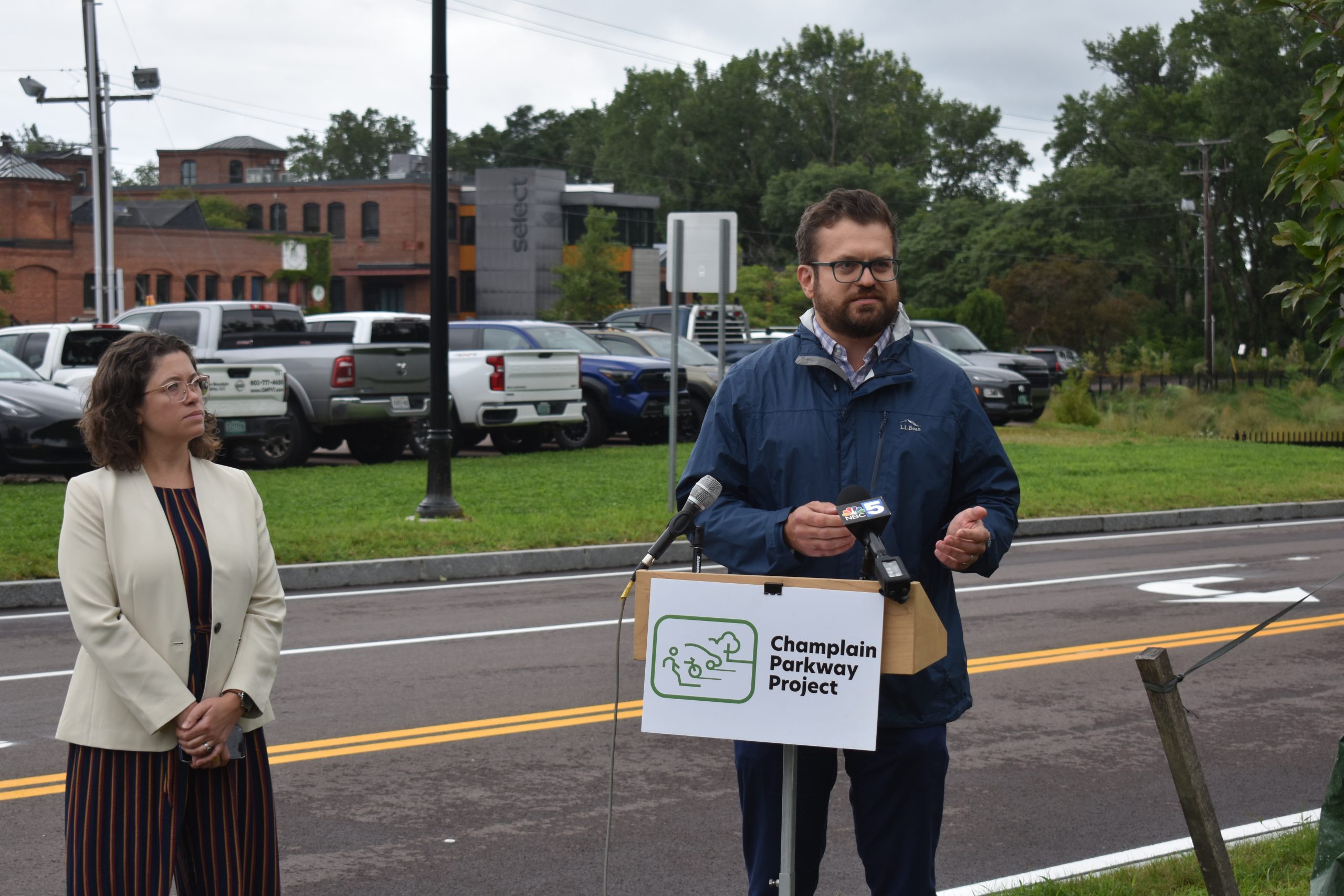 A man speaks at a podium labeled "Champlain Parkway Project" while a woman stands beside him. They are outdoors near a street with parked cars and trees in the background.