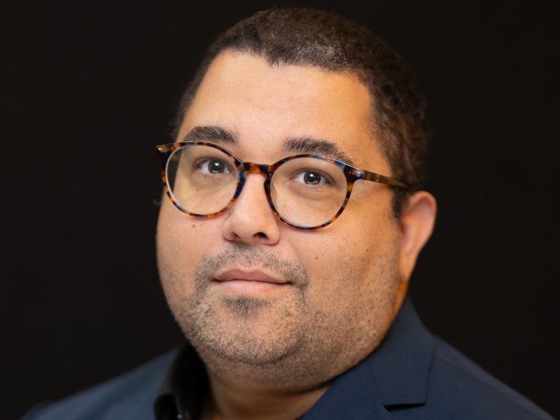 A person with short hair, wearing glasses, and a dark shirt. The background is black.