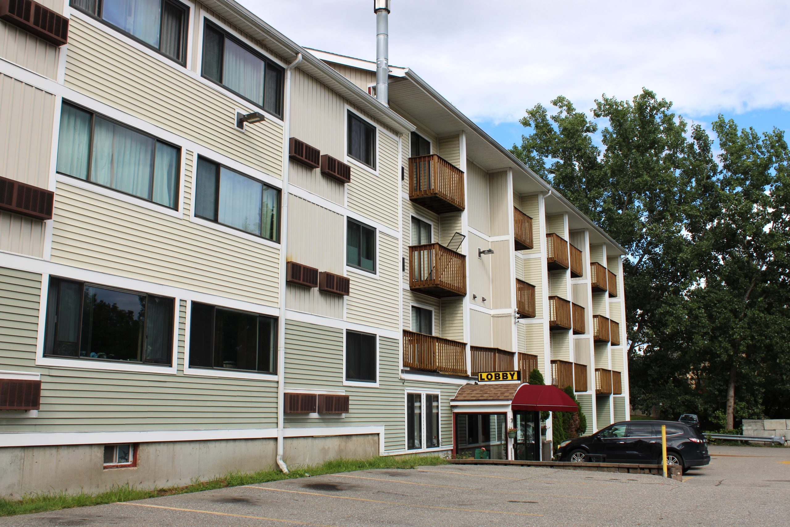 A four-story apartment building with a sign for the lobby. The building has brown balconies and a red awning over the entrance. A black car is parked in front. Large trees are visible nearby.