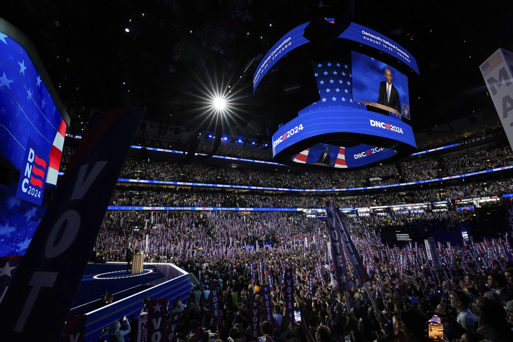 A large crowd in an indoor arena attends a political convention. A speaker stands at a podium on stage, with multiple screens displaying the event and the text "DNC 2024.