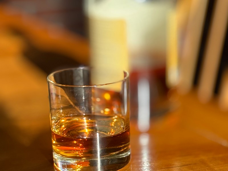 A glass of whiskey on a wooden table with a bottle of whiskey in the background.