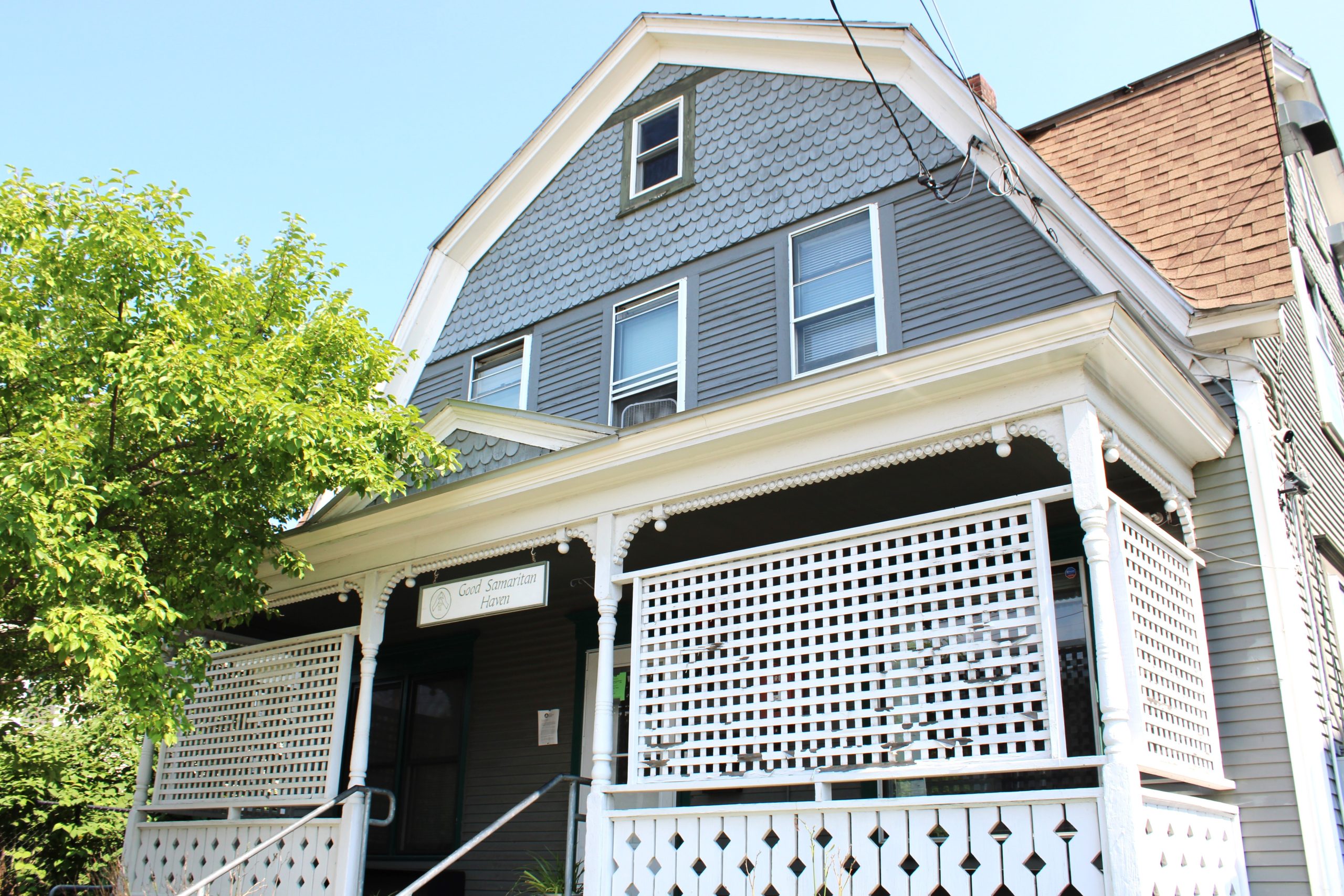 A gray two-story house with white trim features a large front porch with a lattice railing. A tree is on the left side of the house, and there are three windows on the second floor.