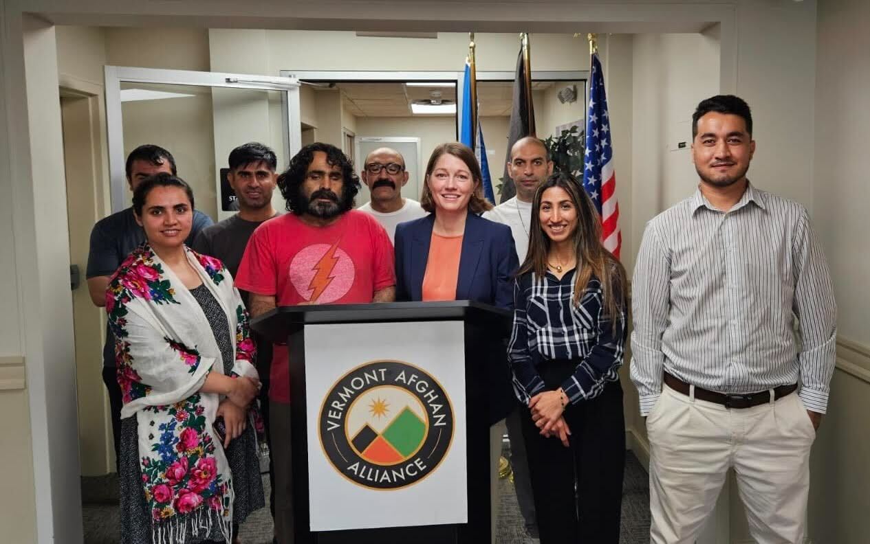 A group of people stands behind a podium with the Vermont Afghan Alliance logo, in front of U.S. and Vermont flags, inside a building.