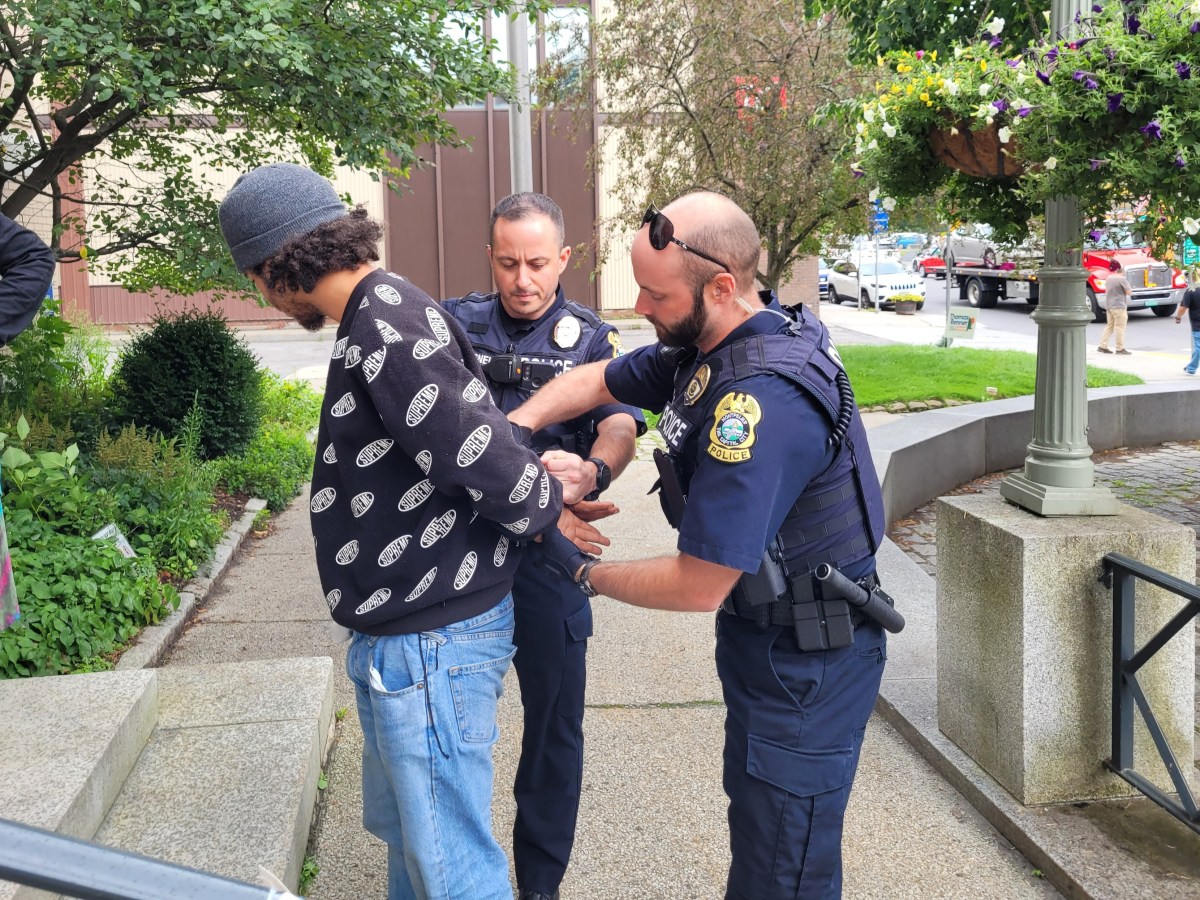 Two police officers are handcuffing a man in a beanie and patterned sweater on a sidewalk, with another officer observing. Trees and parked cars are visible in the background.