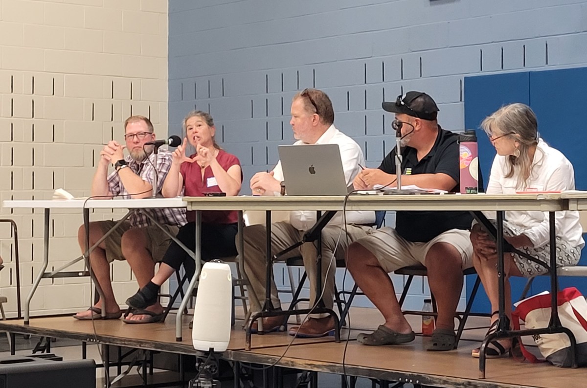 Five people are seated at a long table during an indoor meeting. One person is talking while the others listen. Laptops, microphones, and papers are on the table.