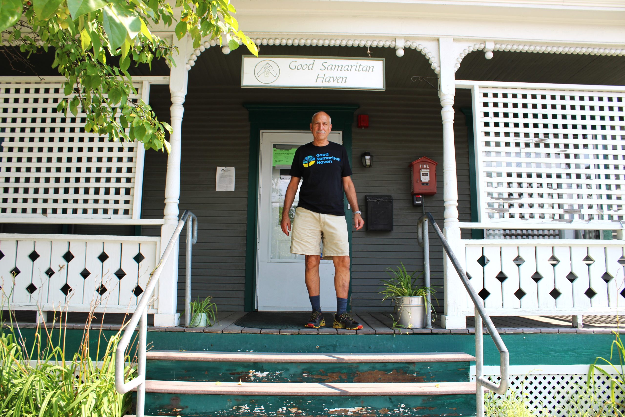 A person stands in front of a building with a sign that reads "Good Samaritan Haven." The porch has green steps, white railings, and potted plants.