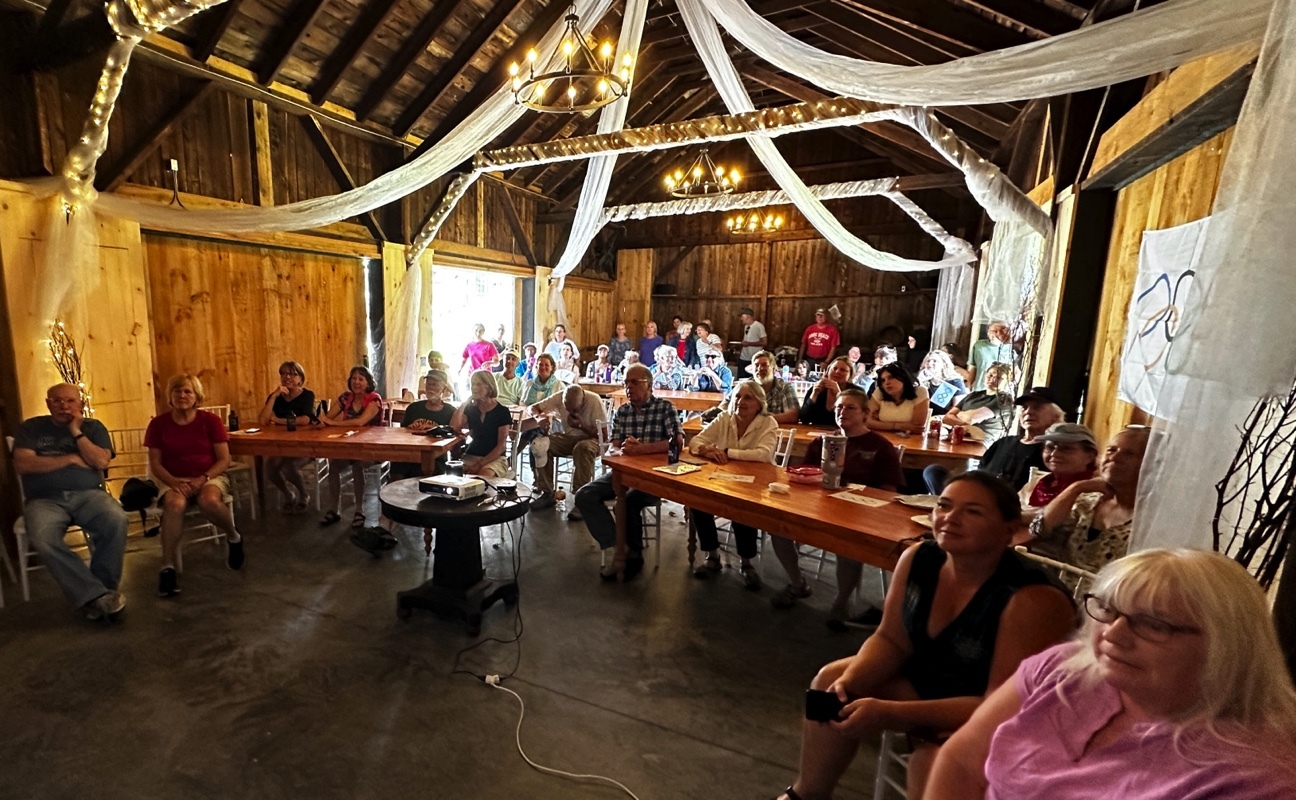 A group of people seated on chairs and benches in a rustic barn decorated with white drapes and string lights, attentively watching a presentation.