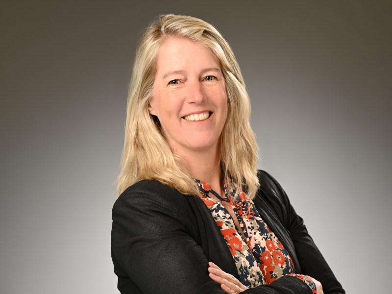 A woman with blonde hair wearing a black jacket and a floral blouse stands smiling with her arms crossed against a plain background.