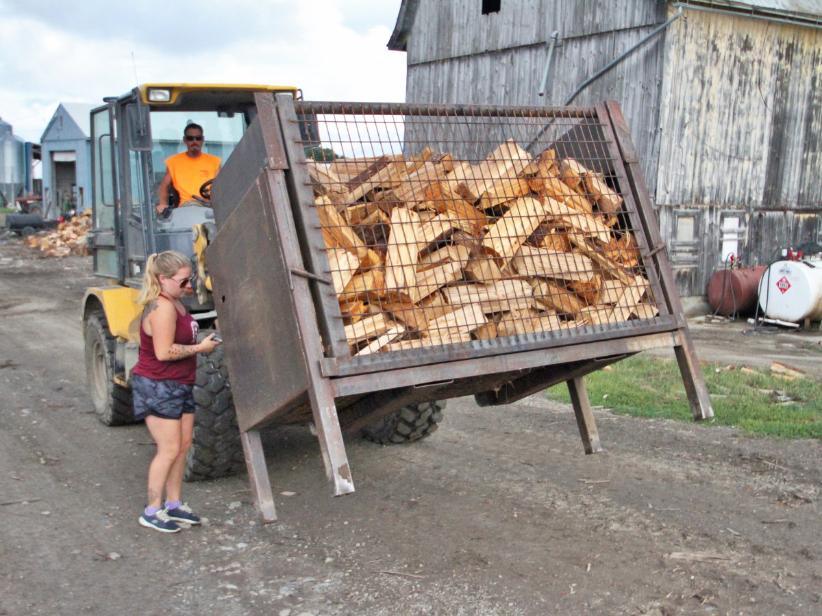 A woman stands next to a loader transporting a large metal bin filled with chopped wood beside a weathered barn.