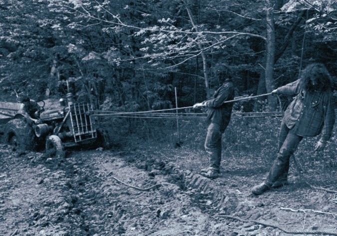 Three individuals pull ropes tied to a vehicle stuck in mud in a wooded area.