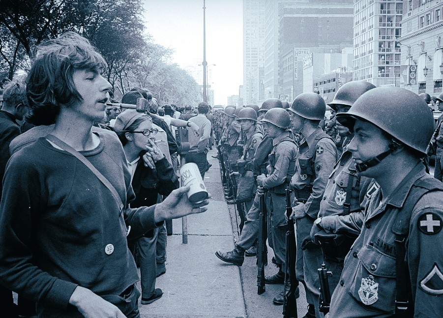 A young man holding a drink stands face-to-face with a line of helmeted police officers during a protest in a city.