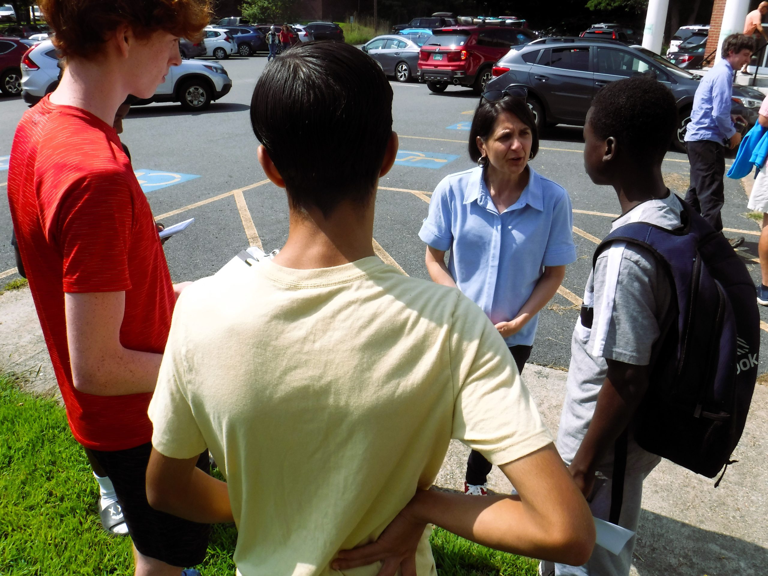 A woman engages in a conversation with three boys at an outdoor location near a parking lot. The boys have backpacks and appear to be listening attentively.
