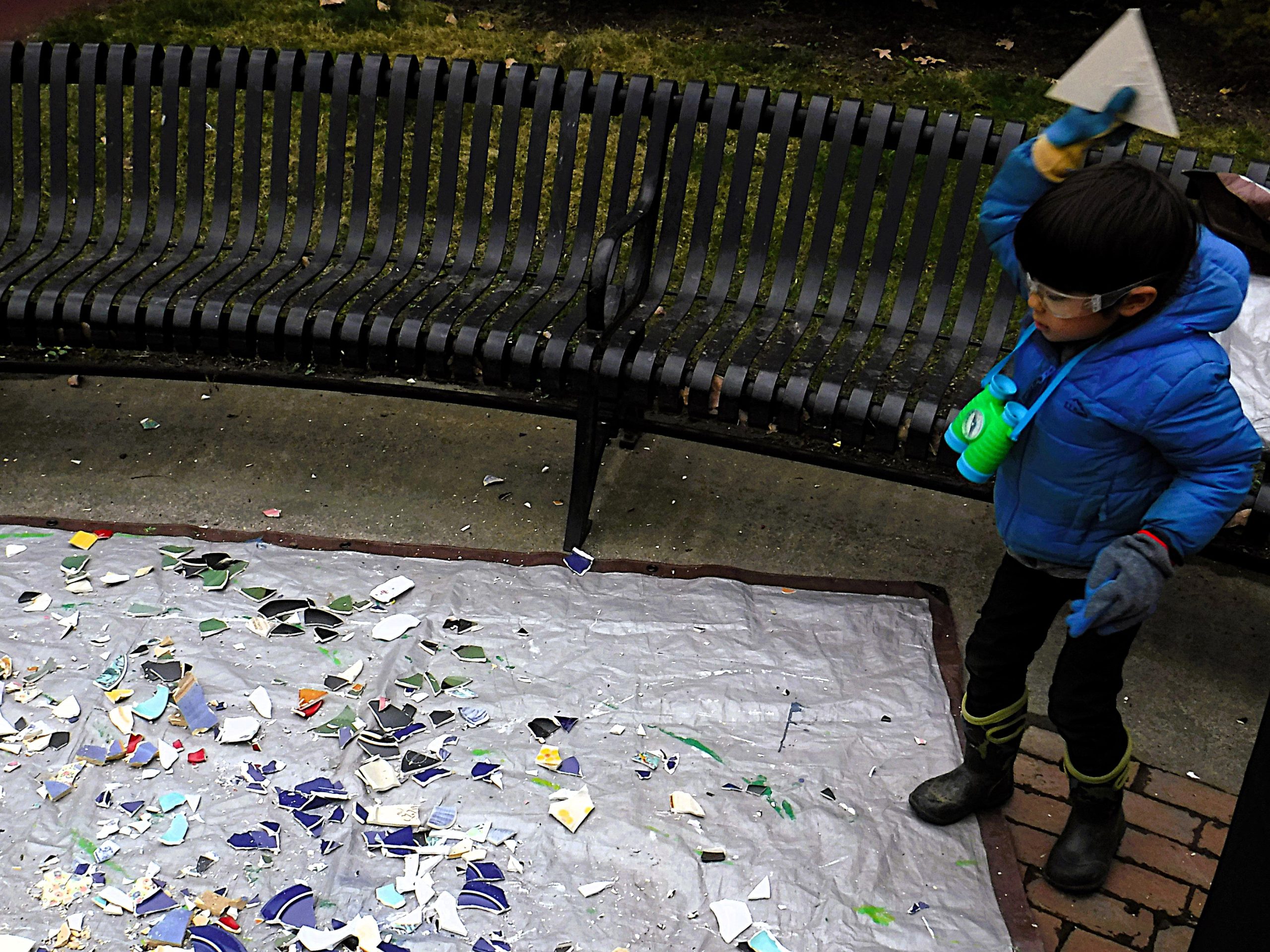 A child wearing goggles and gloves prepares to break a piece of tile over a tarp covered with tile fragments in a park, near a curved bench.
