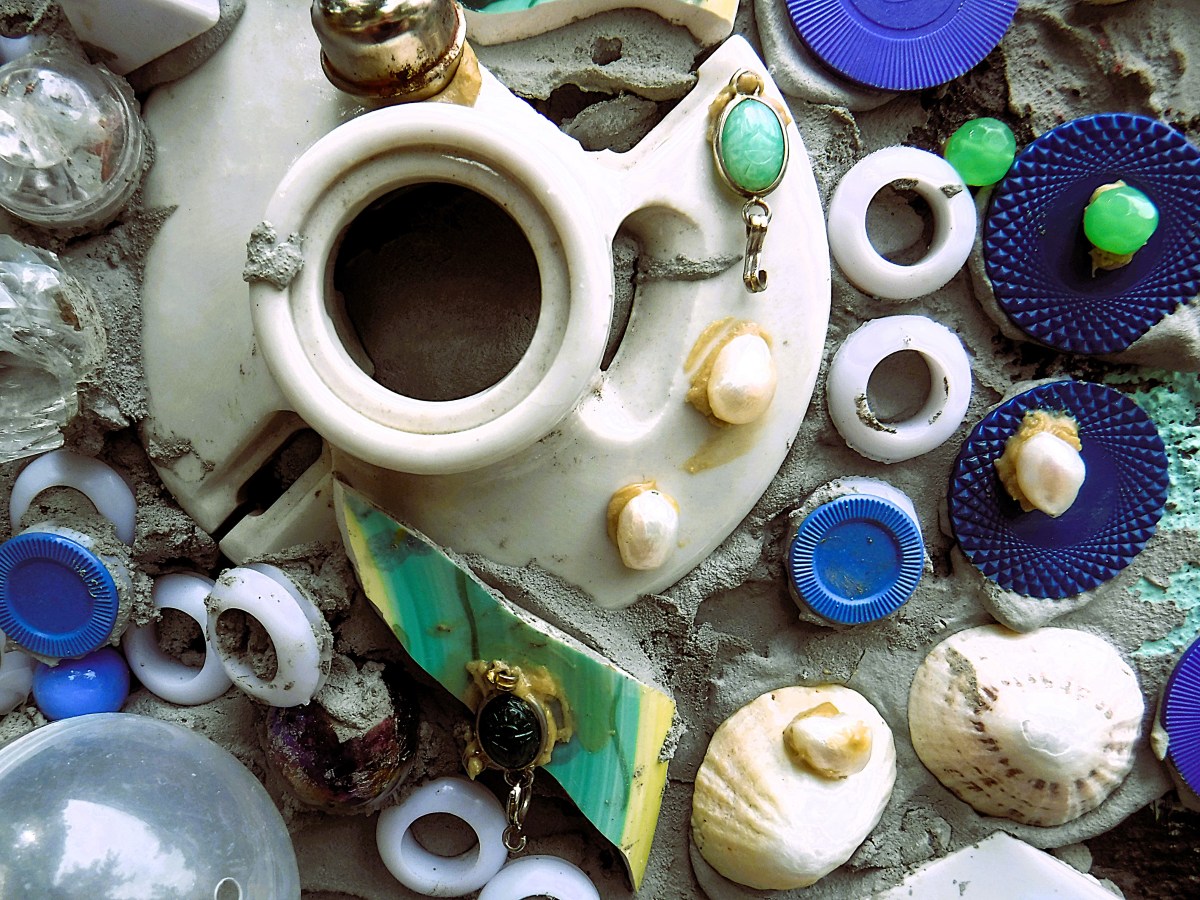 A close-up of various objects embedded in cement, including ceramic fragments, plastic caps, shells, and washers, creating a textured mosaic.