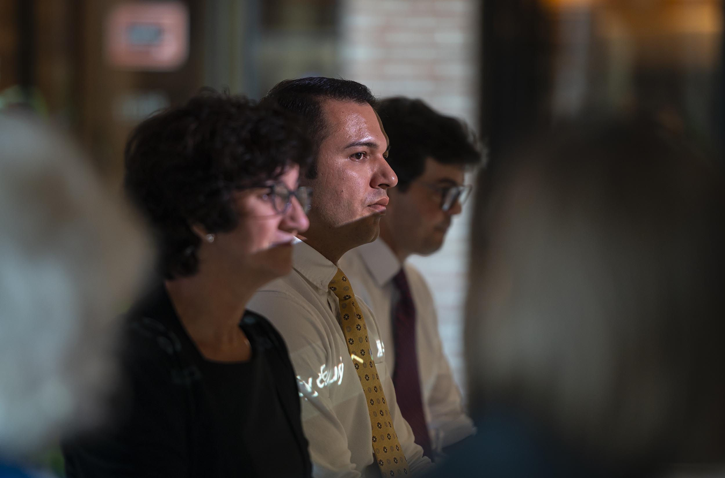 A group of people sitting attentively indoors; two men are wearing glasses, and a man in the center is wearing a white shirt with a yellow tie. The focus is on the man in the center.