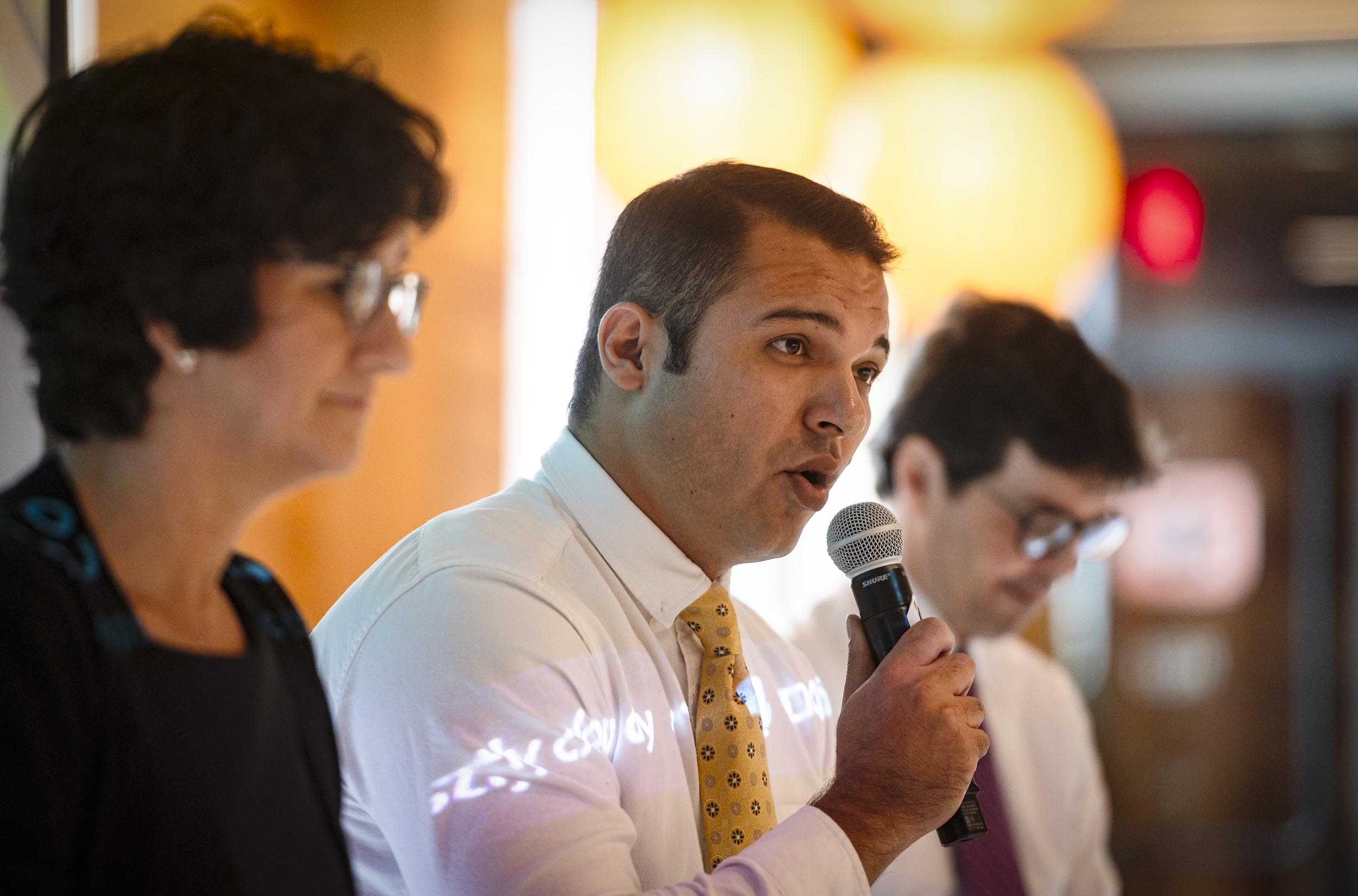 Three individuals are standing together, one of them holding a microphone and speaking, while the others listen attentively. They appear to be in a professional setting.