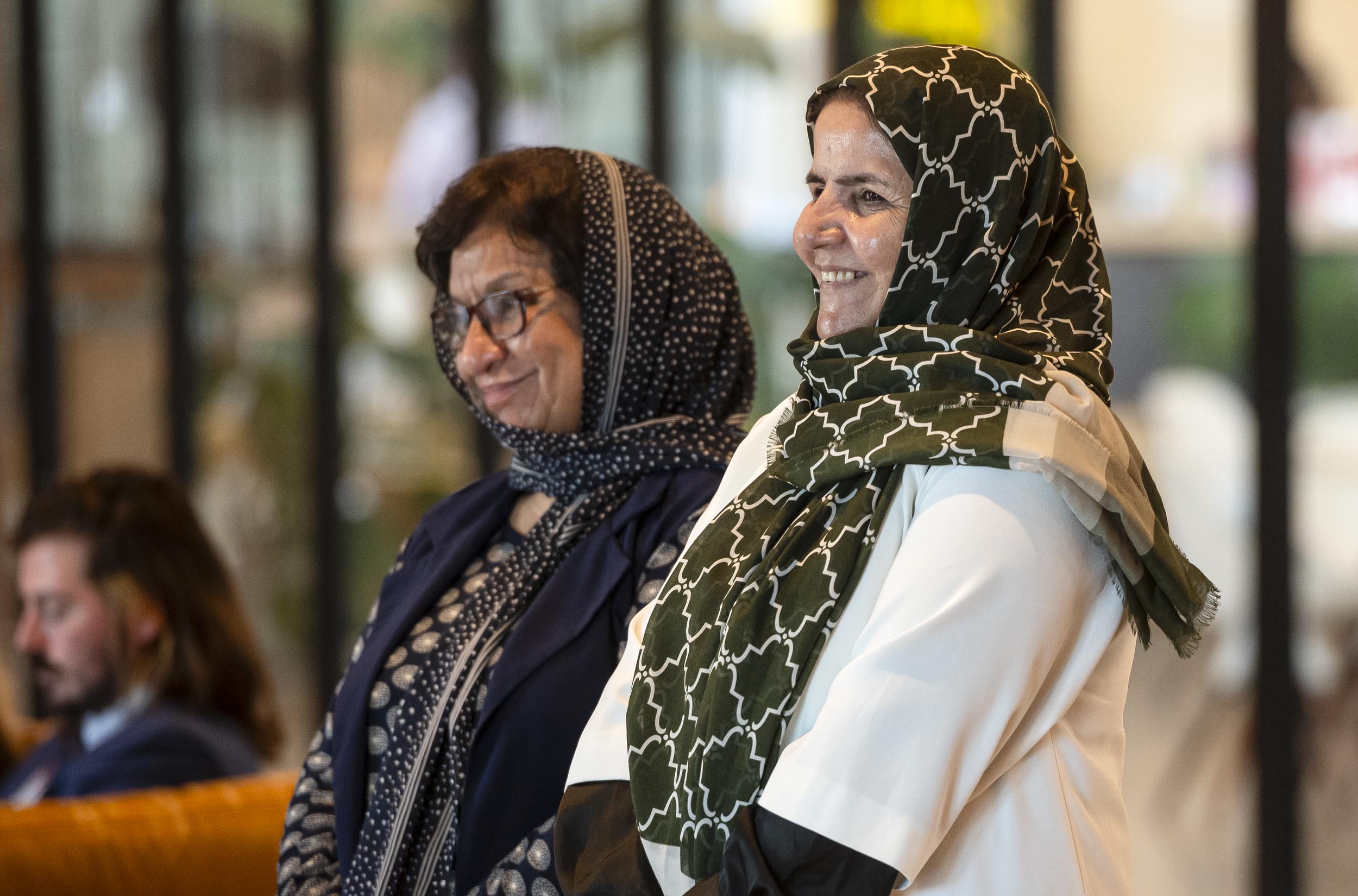 Two women wearing patterned headscarves and smiling stand indoors with glass windows in the background.