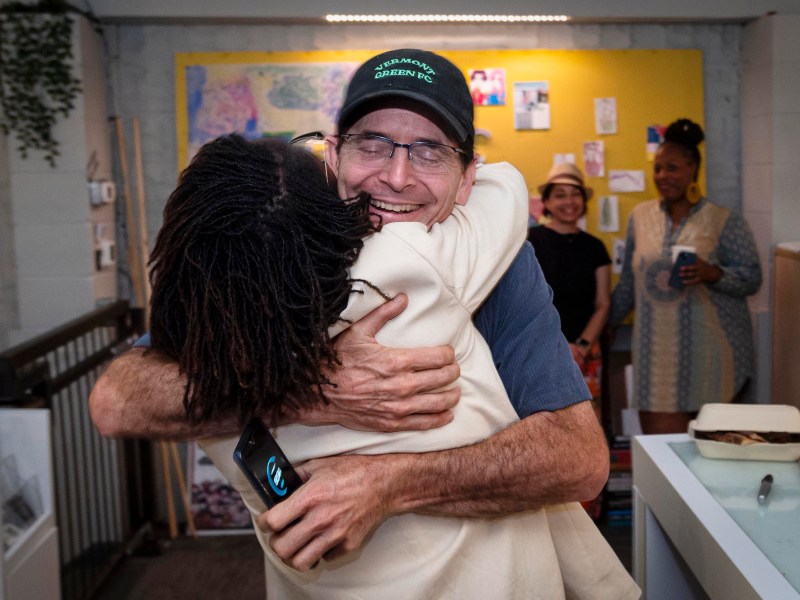 Two people embrace in a warm hug in an indoor setting. One individual holds a smartphone, while others in the background appear to be smiling and conversing.