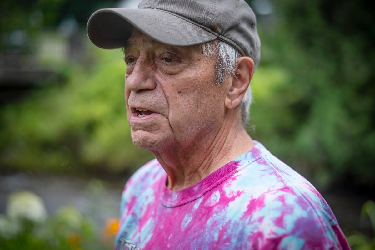 An elderly man in a tie-dye shirt and cap stands outdoors, looking to the side with greenery in the background.