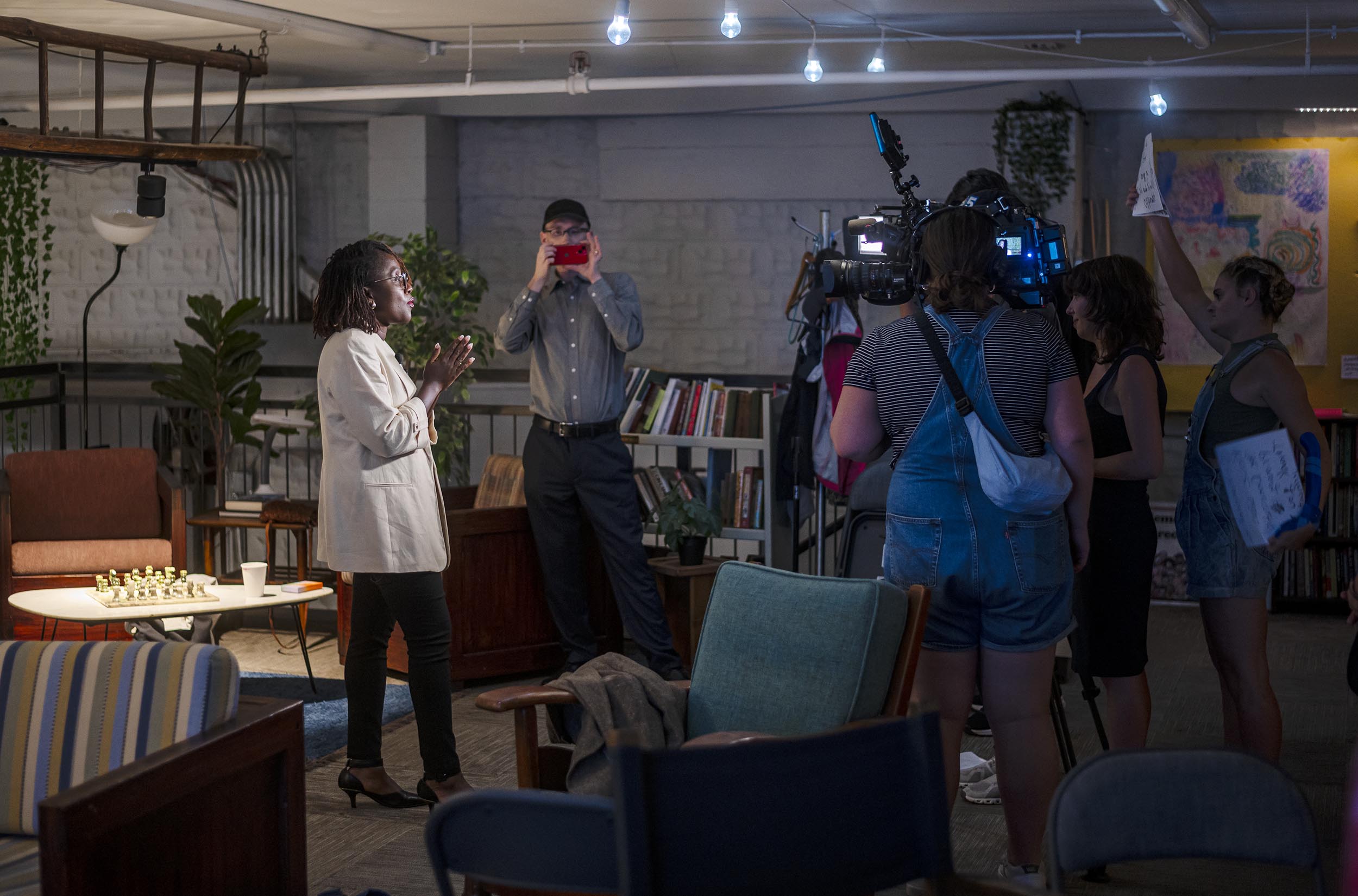 A person is speaking in front of a camera crew in a furnished indoor setting with bookshelves and seating areas in the background.