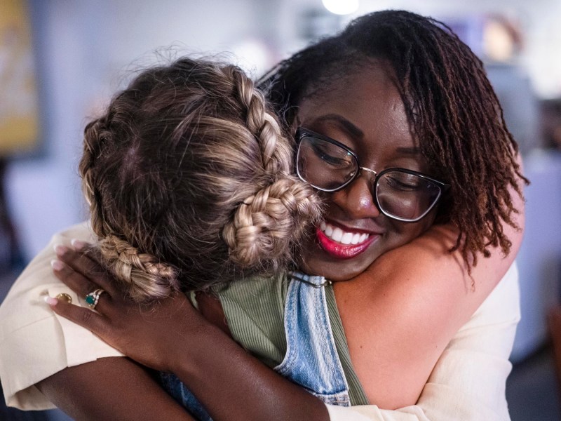 Two people embracing closely, one with braided hair and the other with dreadlocks and glasses, both smiling warmly.