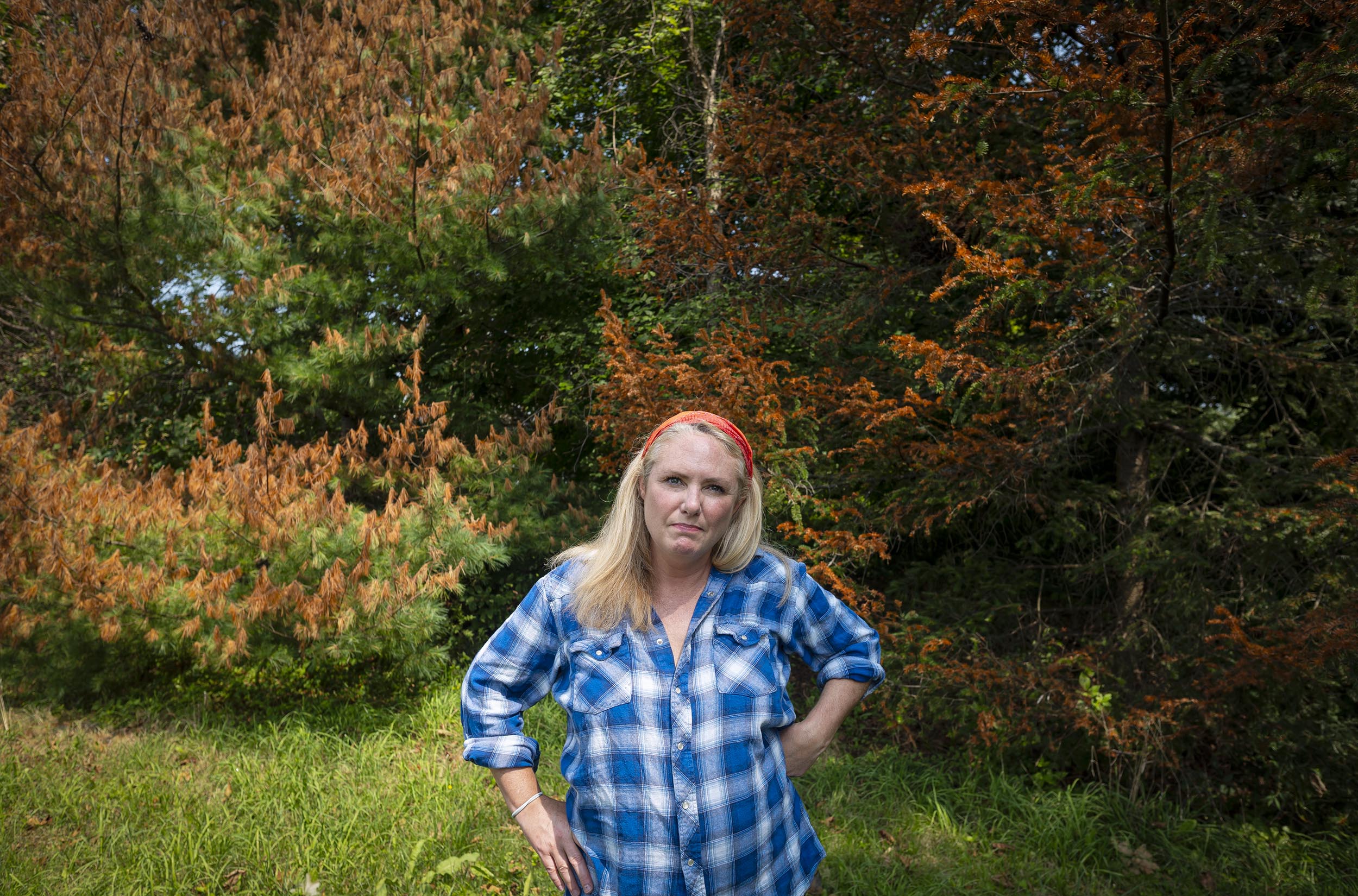 A woman wearing a blue plaid shirt and red headband stands with hands on hips in front of trees with green and orange foliage.