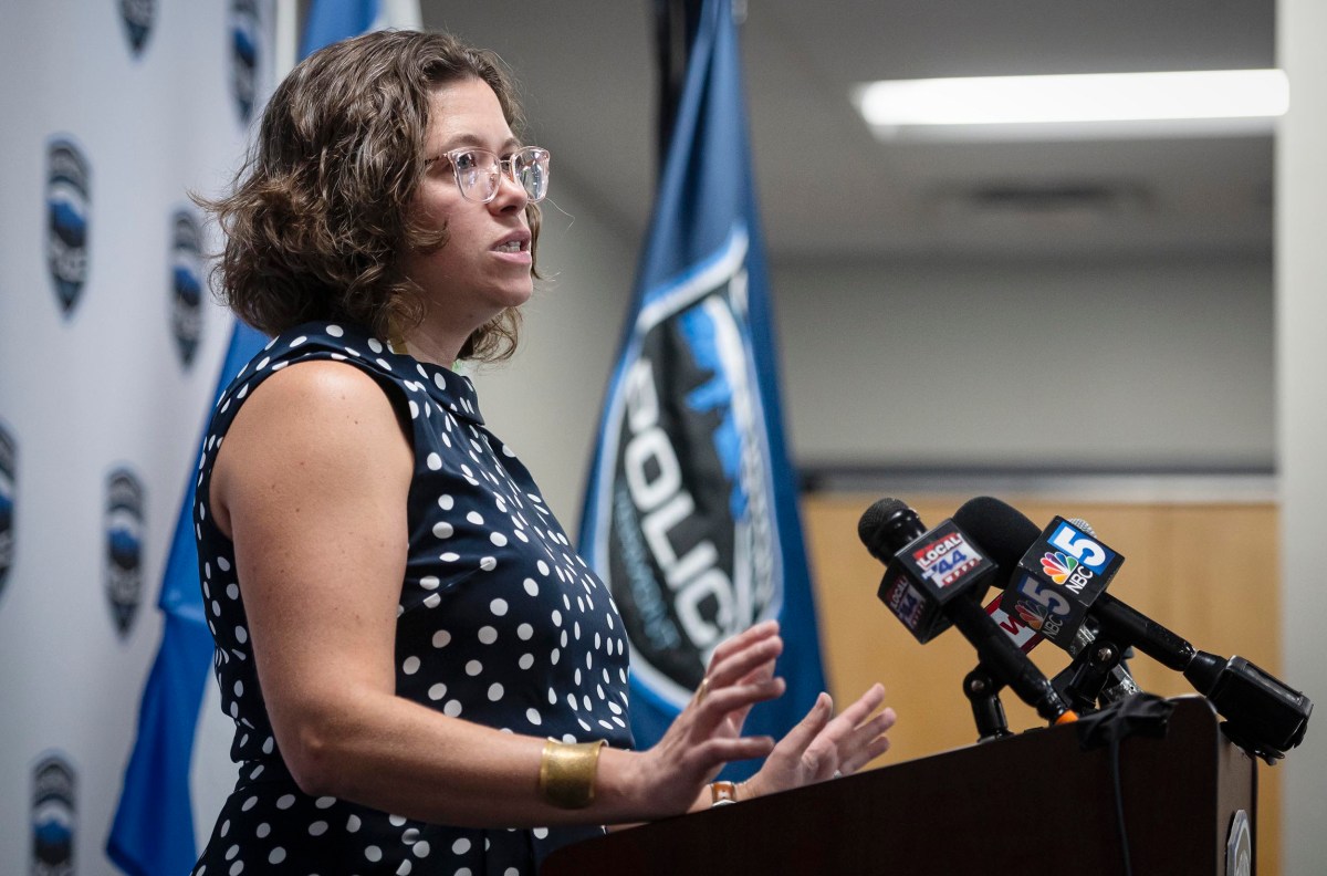 A woman in a polka dot dress speaks at a podium with multiple microphones. Police department insignia and flags are visible in the background.
