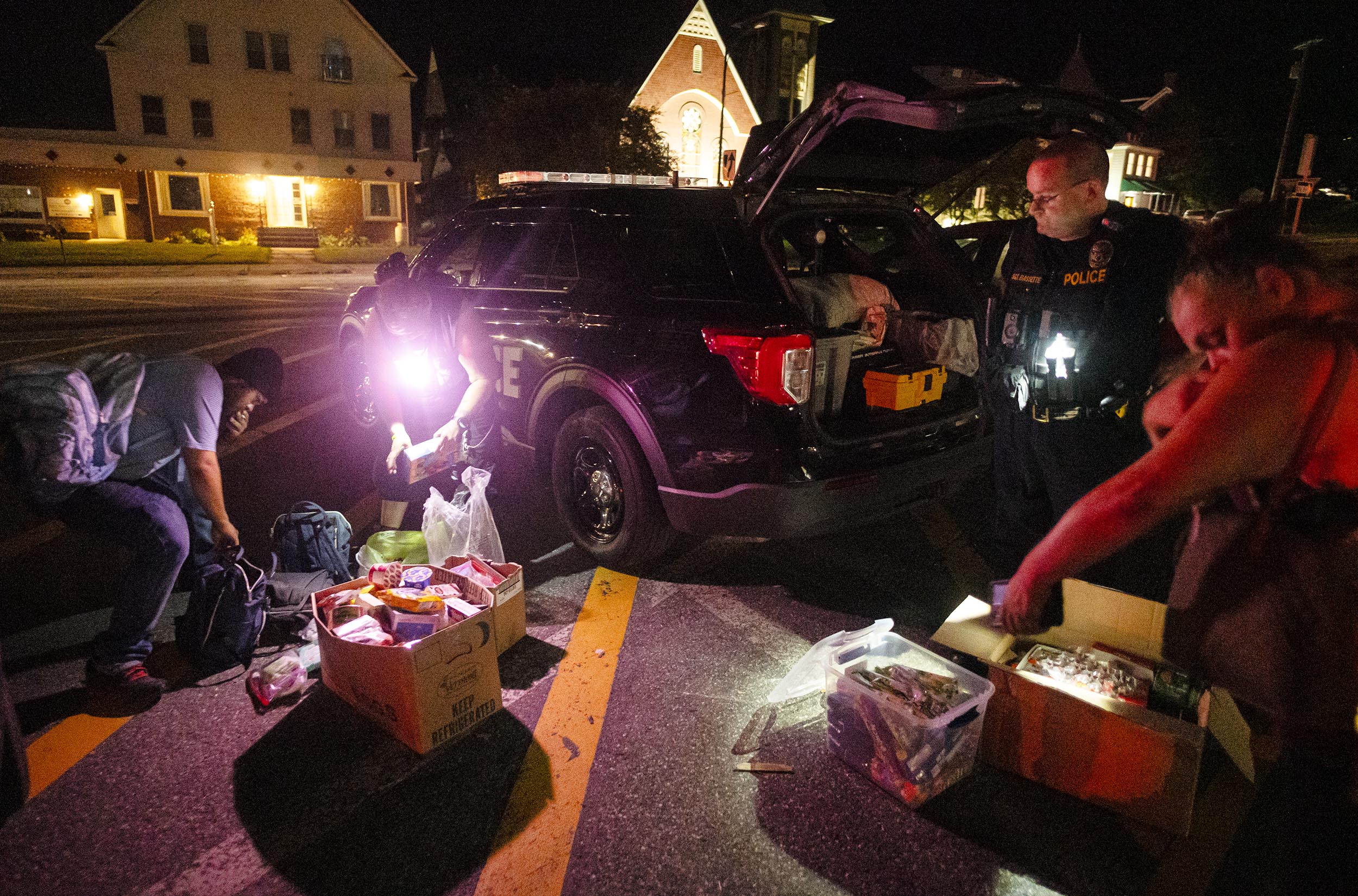 Police officers and civilians at night by an open police SUV, inspecting boxes and bags of items on the ground under streetlights.