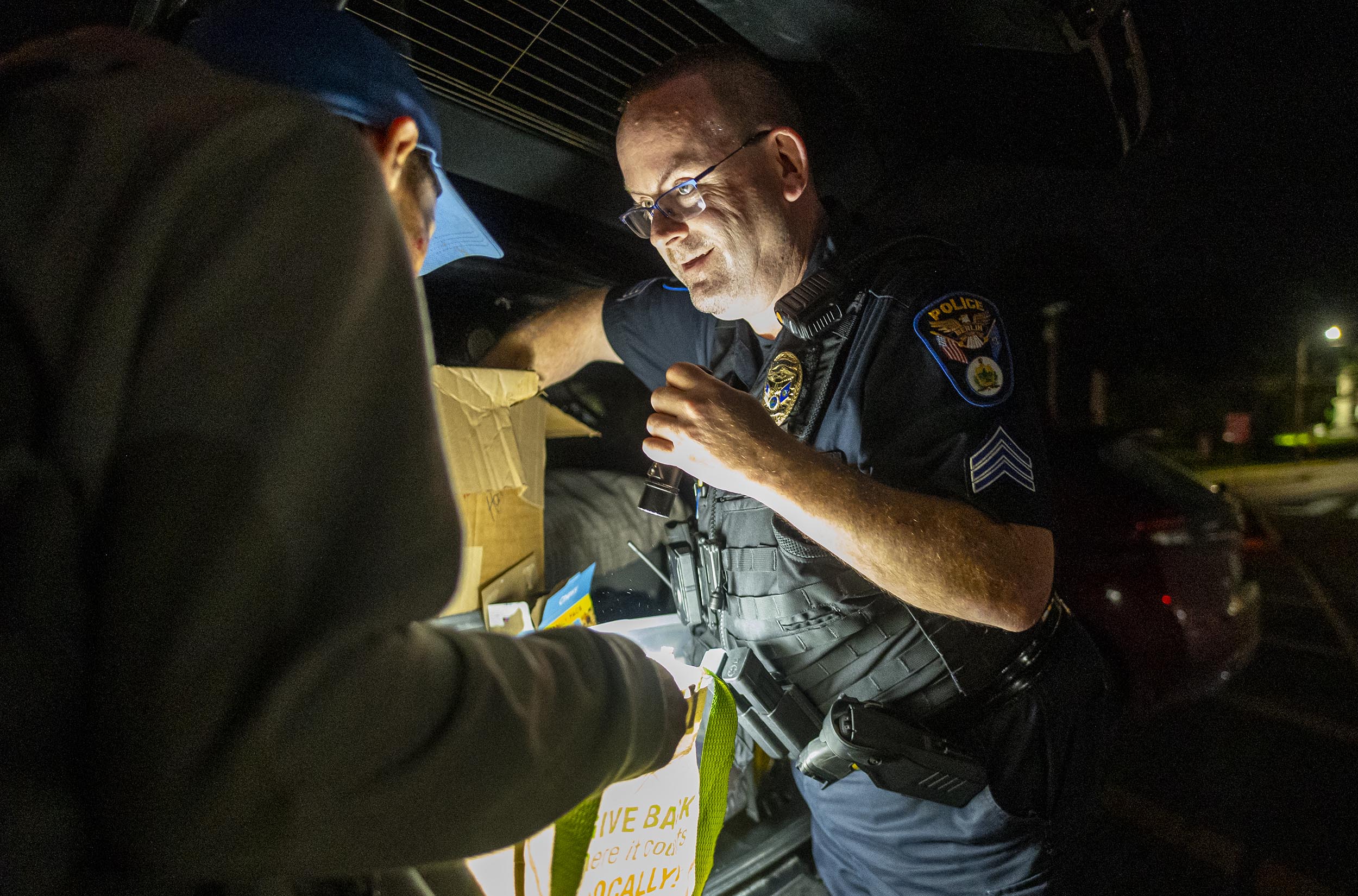 A police officer in uniform speaks with a person wearing a cap in front of an open car trunk at night, illuminated by a light source.