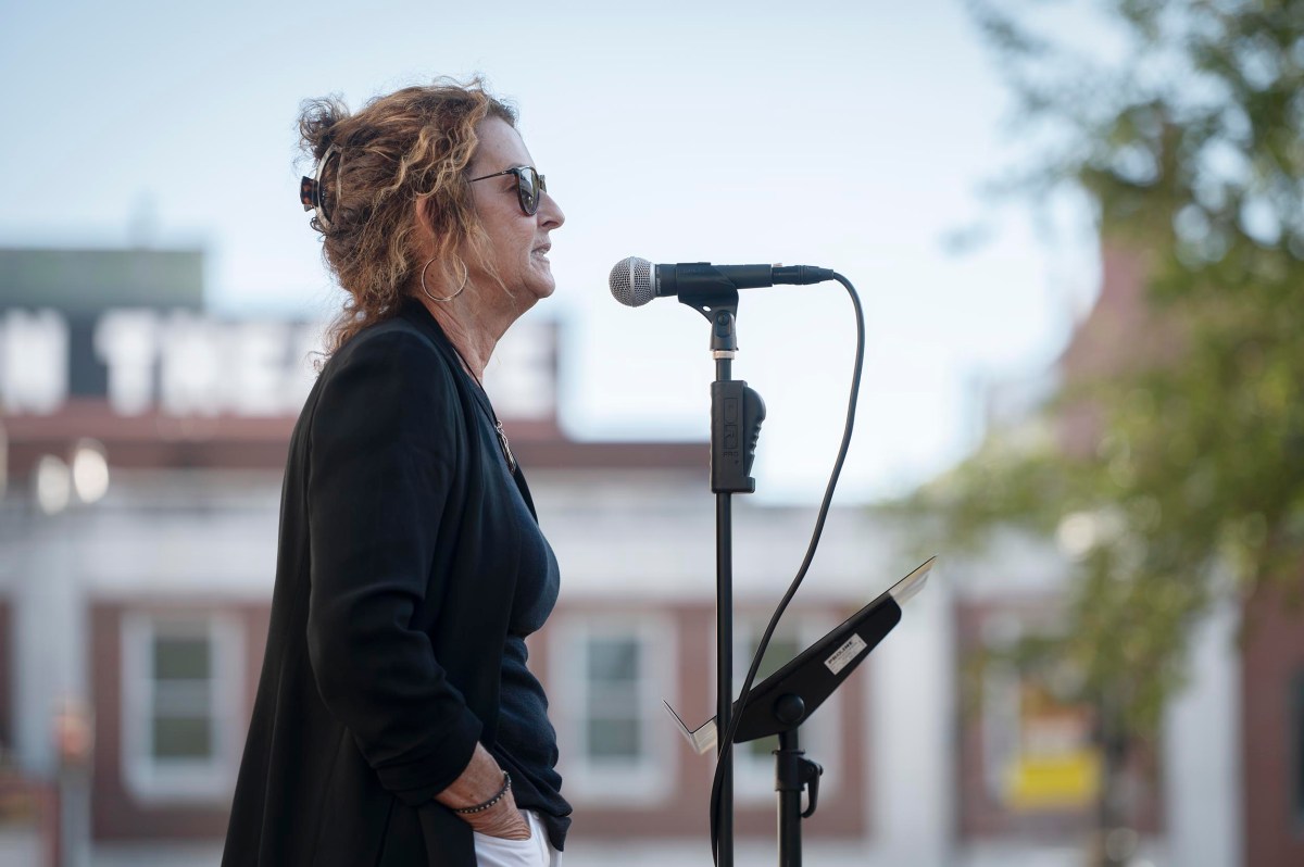 A person with curly hair and sunglasses speaks into a microphone at an outdoor event, with a building and some trees in the background.