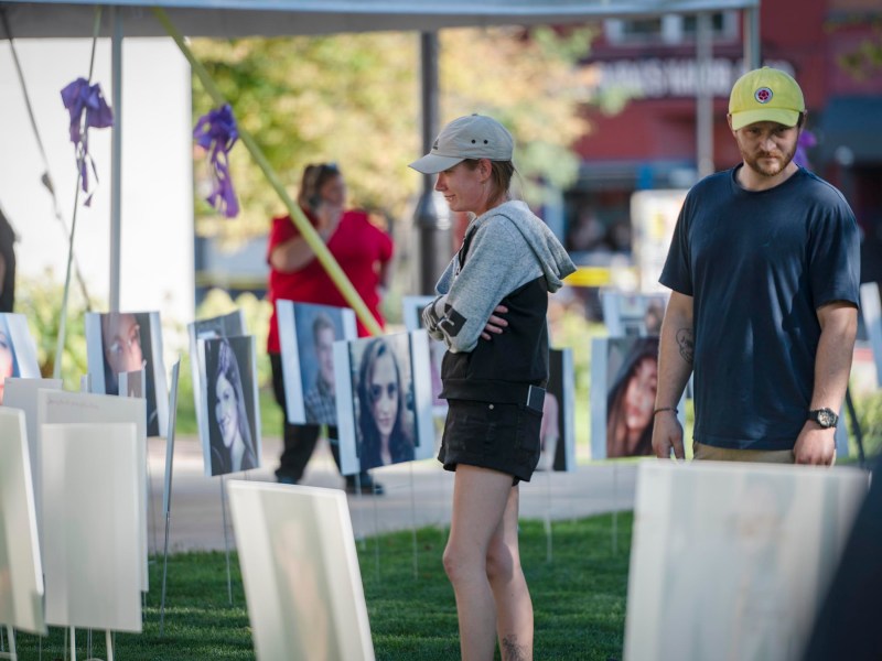 People at an outdoor photo exhibit featuring portraits on display under a tent. A woman in a cap and shorts and a man in a baseball cap are looking at the photos.