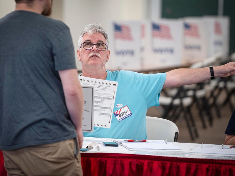 A man in a blue shirt and glasses holds a ballot while speaking to another person at a voting station with American flags in the background.