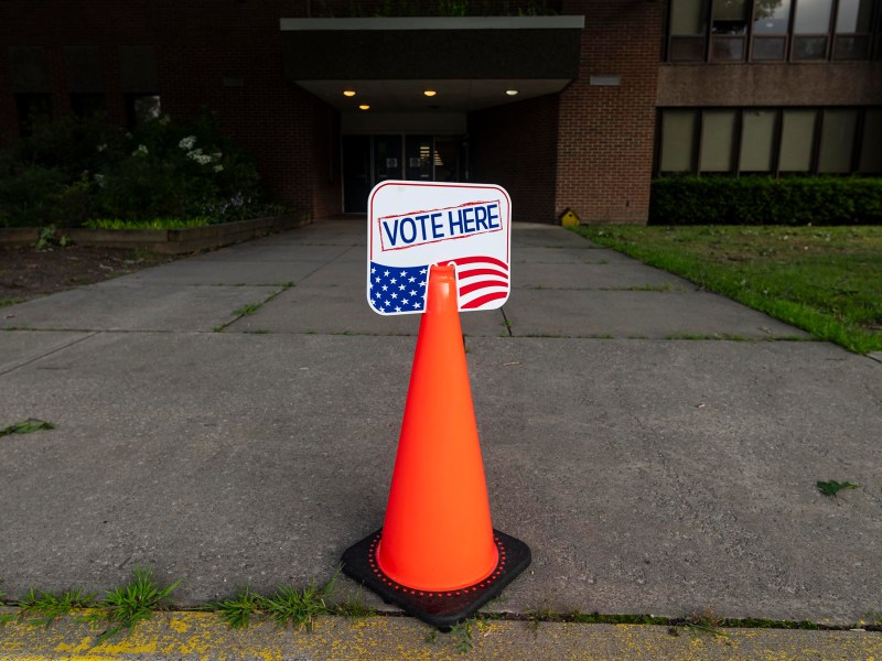 Orange traffic cone with a "Vote Here" sign placed in front of a building entrance, indicating it is a polling or voting station.