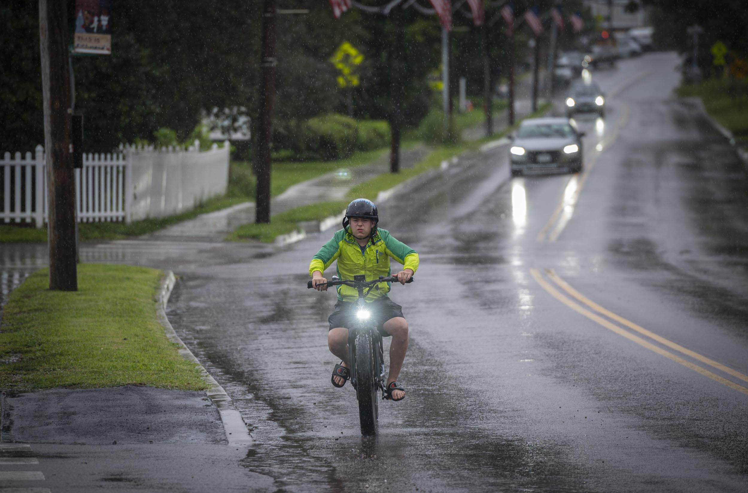 A cyclist wearing a green jacket and helmet rides through the rain on a wet road. Cars are visible in the background driving on the same road.
