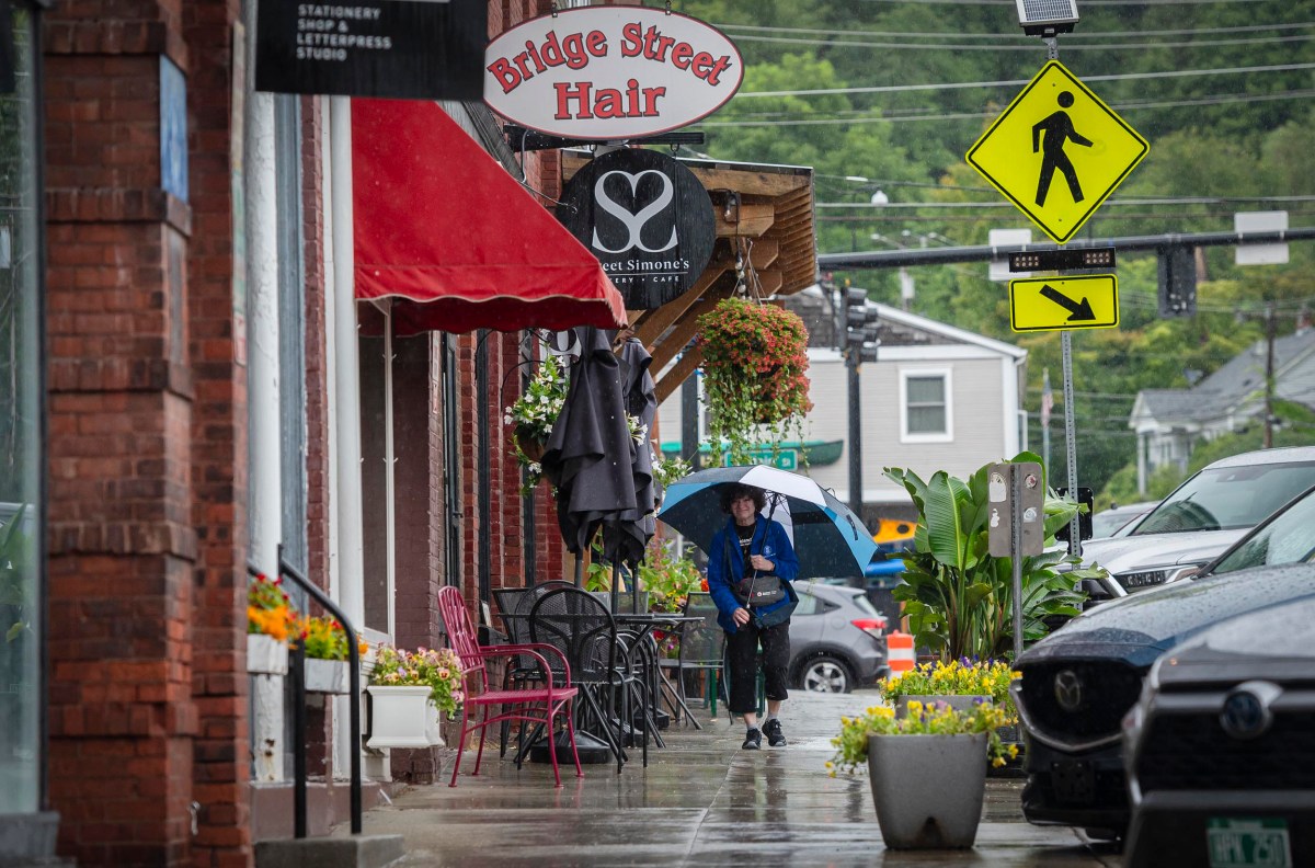 A person with an umbrella walks along a rainy street lined with shops and parked cars. Signs for "Bridge Street Hair" and a pedestrian crossing are visible.