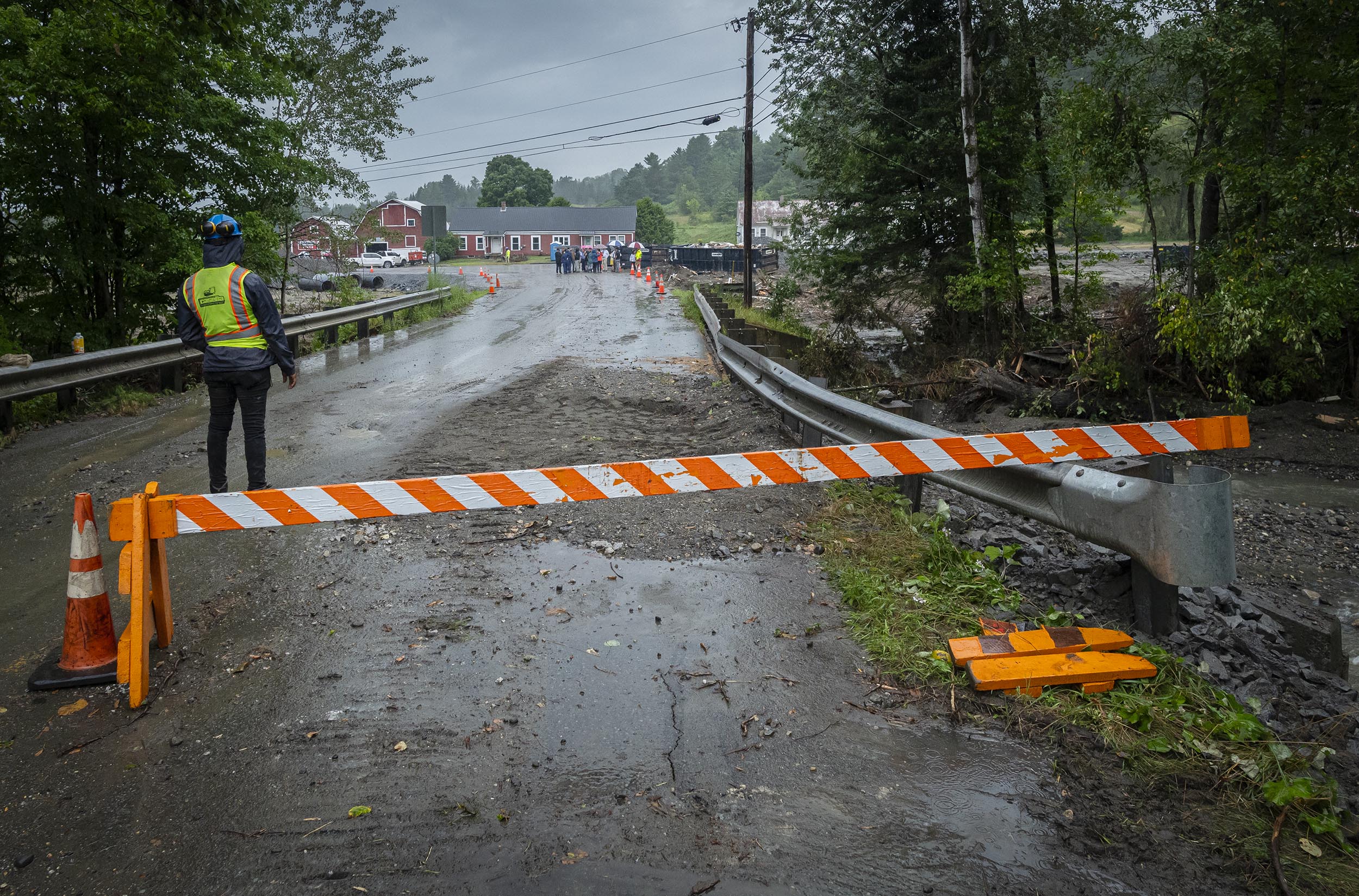 Worker in safety gear stands near a road barricaded with orange and white striped barrier, debris scattered on the road, with people and buildings visible in the background.