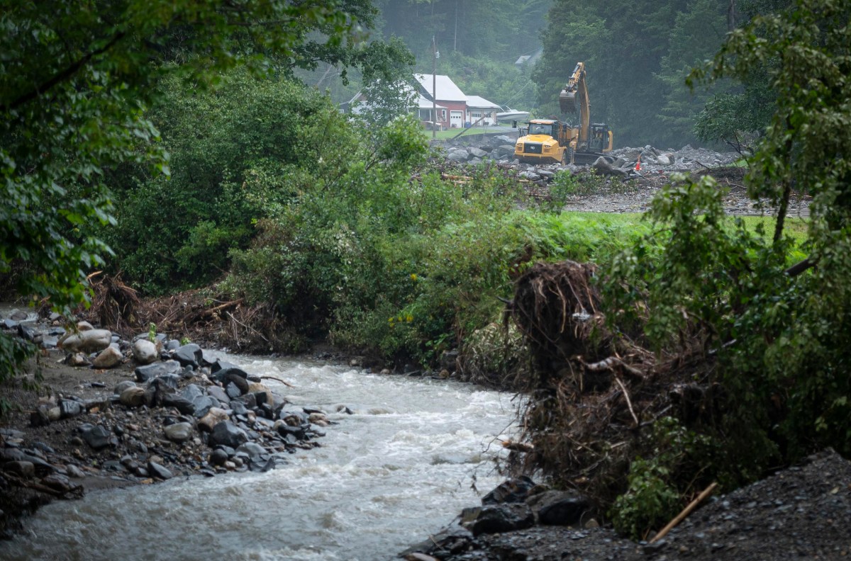 A yellow excavator works on a construction site near a river, surrounded by trees and greenery, with a house visible in the background.