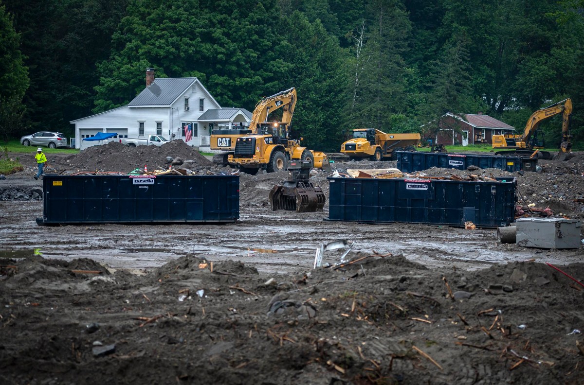 Construction site with heavy machinery, including excavators and loaders, near residential houses. Large dumpsters contain debris, and the ground is muddy. Trees are in the background.