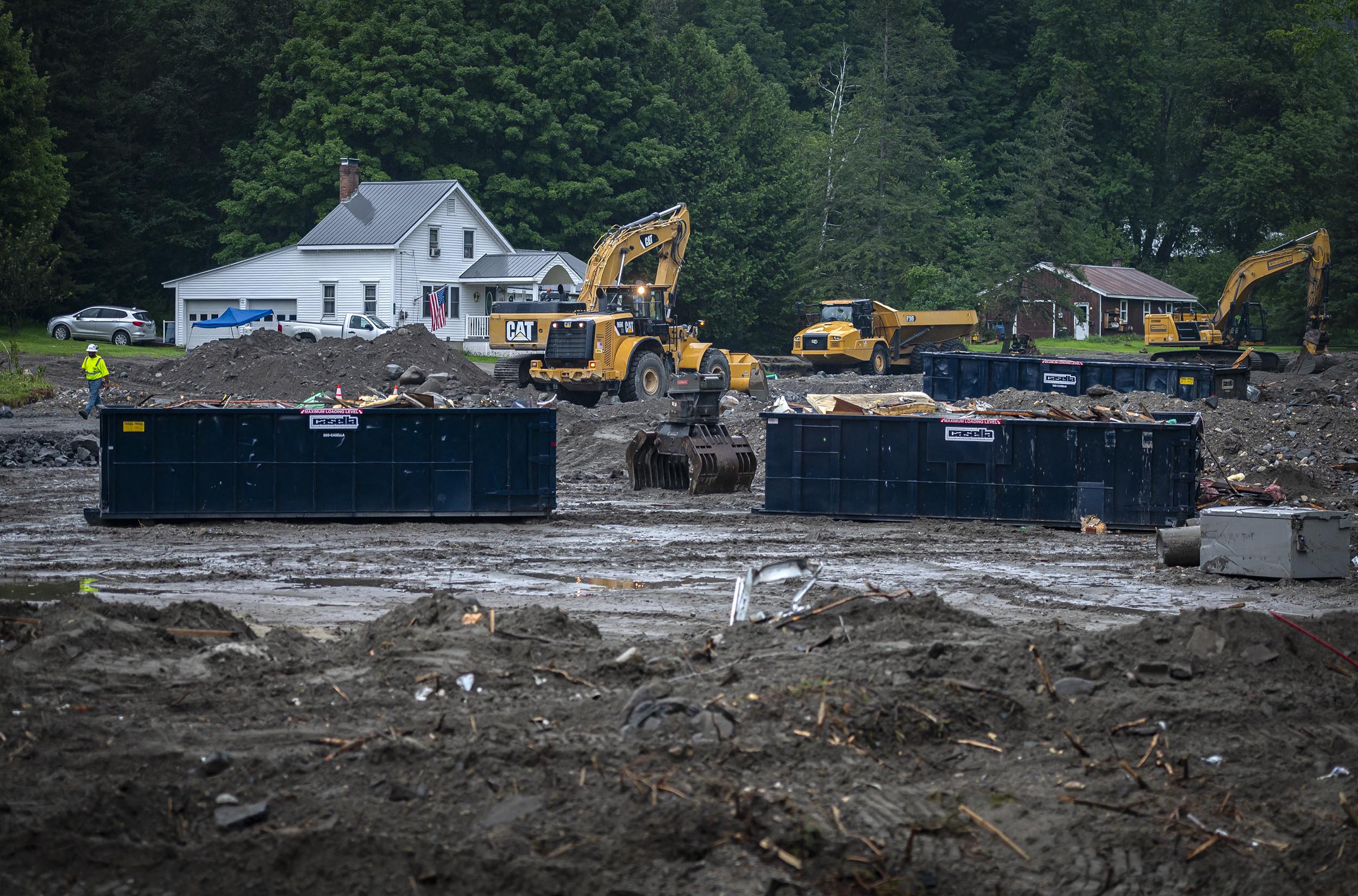 Construction site with heavy machinery, including excavators and loaders, near residential houses. Large dumpsters contain debris, and the ground is muddy. Trees are in the background.
