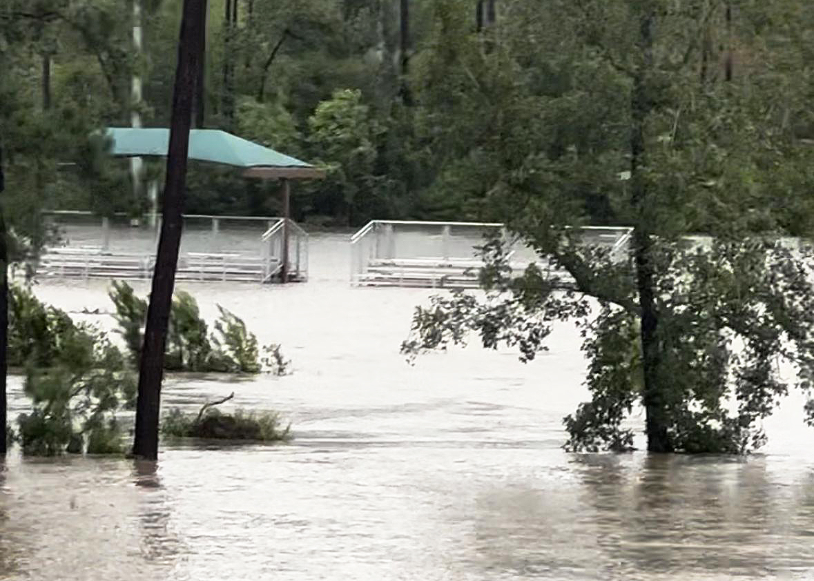 A flooded area with submerged bleachers and trees. A pavilion with a teal roof is visible in the background amidst rising water levels.