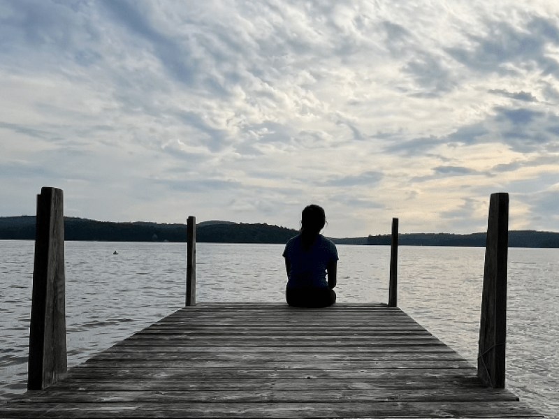 A person sits at the end of a wooden dock, facing a tranquil lake under a cloudy sky.