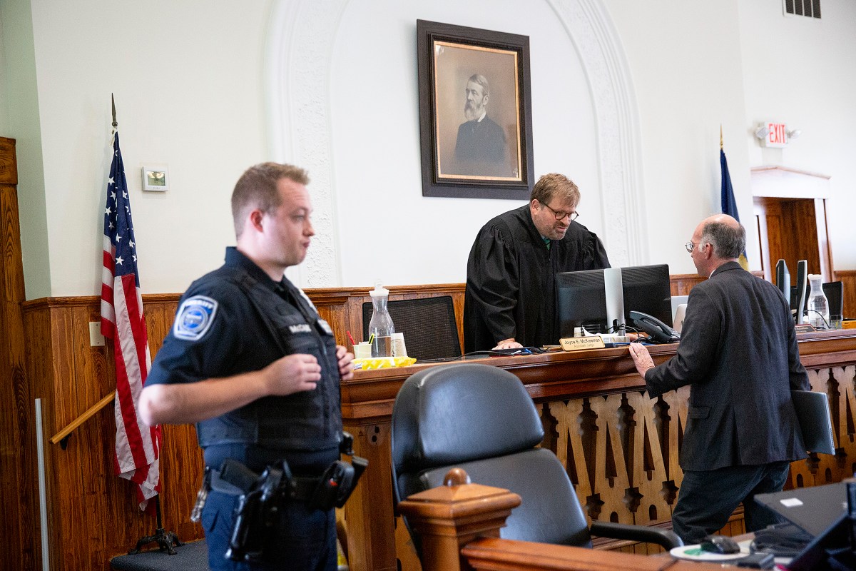 A judge in a black robe speaks to a lawyer at the bench in a courtroom, while a police officer stands nearby. An American flag and a portrait hang on the wall in the background.