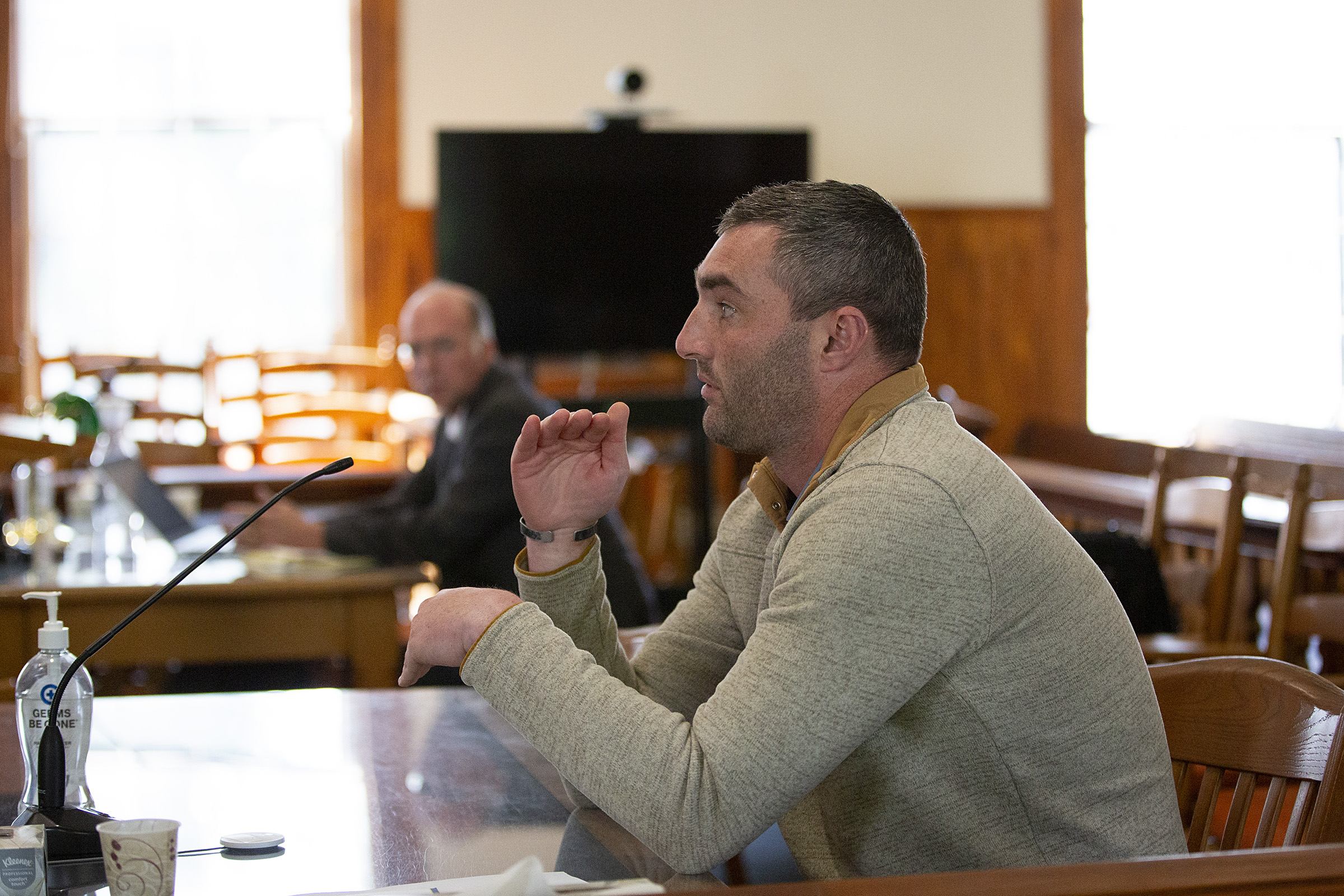 A man speaking into a microphone is seated at a table in a room with wooden chairs and a television screen in the background. Another man is seated at a separate table using a laptop.