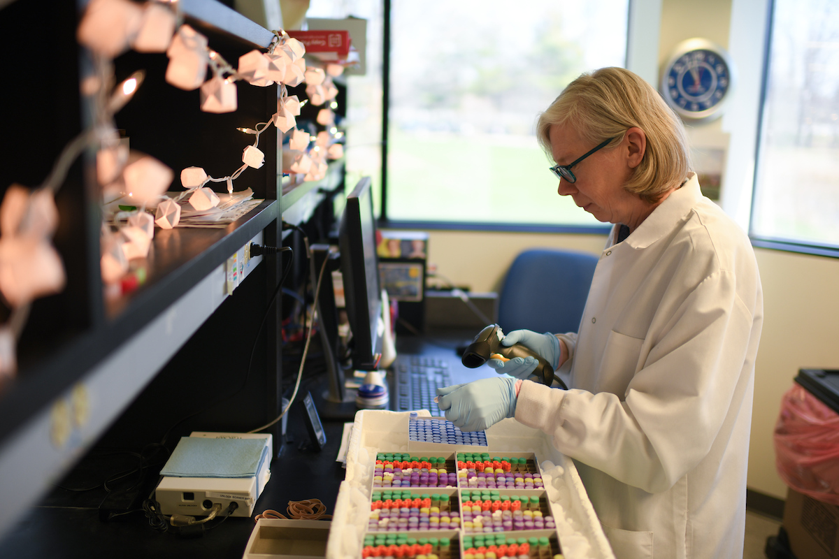 A person in a lab coat and gloves works with samples in a laboratory, scanning barcodes on colorful vials. Shelves above are decorated with string lights, and a clock is on the wall.