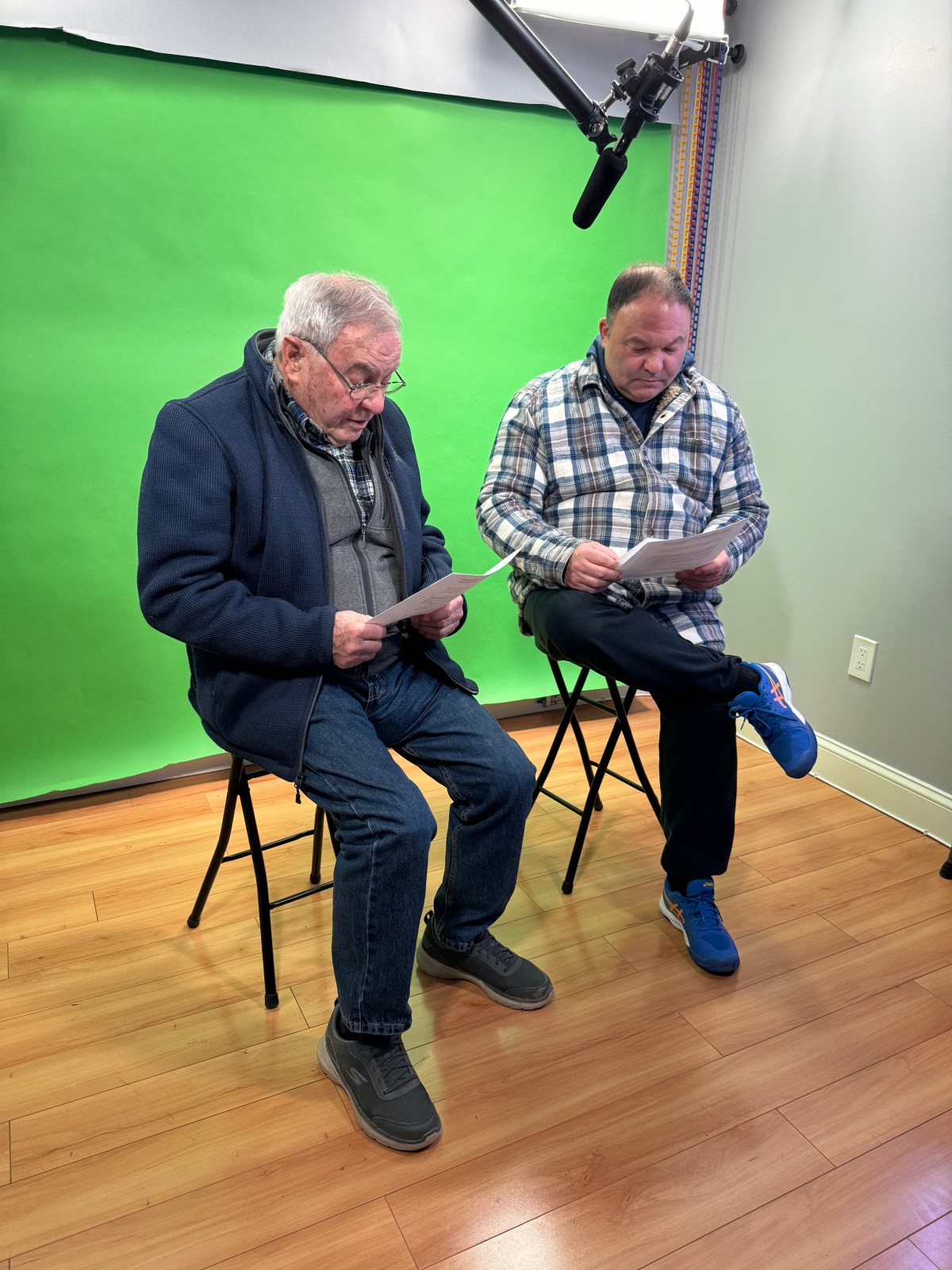 Two men are sitting on stools in front of a green screen, reading papers. A microphone is visible above them.