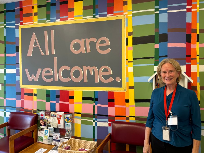 A person stands smiling next to a colorful wall with a sign that reads "All are welcome." The area features chairs, a pamphlet stand, and a basket on a table.
