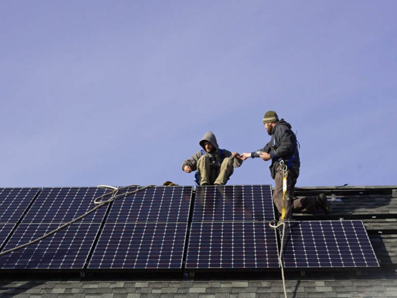 Two workers on a roof installing solar panels, one handing a tool to the other.